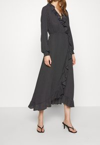 Svart prickig klänning med volang vid halsringning och fåll, långa ärmar och omlottdesign. Tillverkad av lättviktsmaterial.