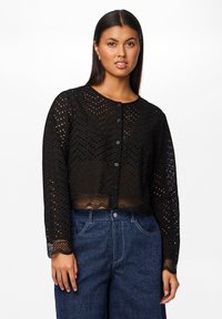 Cardigan corto nero con un design a occhielli testurizzato, maniche lunghe, scollatura rotonda e cinque bottoni sul davanti. Dettaglio dell'orlo ondulato.