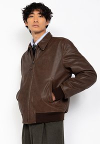 Chaqueta de cuero marrón con un cuello clásico, bolsillos frontales y puños acanalados. Textura suave y diseño minimalista.