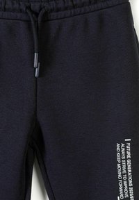 Gros plan sur un pantalon de survêtement noir avec une taille à cordon et un texte blanc sur la jambe droite indiquant "Générations Futures 2025. Toujours s'efforcer de s'améliorer et de continuer à avancer."