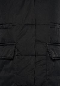 Czarna pikowana kamizelka z dwoma przednimi kieszeniami, gładką fakturą i poziomymi przeszyciami. Materiał wydaje się trwały i odporny na warunki atmosferyczne.