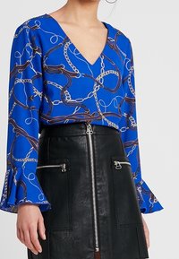 Modrá blůza s výstřihem do V, zdobená potiskem řetězce, v kombinaci s černou koženou sukní s předním zipem a kapsami.