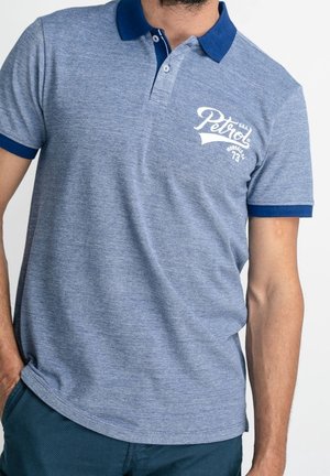 Mężczyzna ubrany w niebieski melanżowy polo z granatowym kołnierzem i wykończeniami rękawów, z logo "Petrol" na piersi oraz niebieskie teksturowane spodnie.