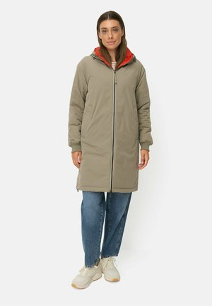 Vrouw draagt een lange beige capuchonjas met rode voering, blauwe spijkerbroek en witte sneakers, staand voor een effen achtergrond.