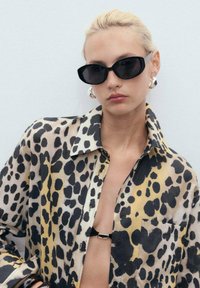 Camicia con stampa leopardata in tessuto trasparente presenta colori nero, giallo e beige, con una chiusura davanti a bottoni e una tasca sul petto. Occhiali da sole neri e orecchini audaci.
