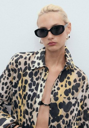 Camicia con stampa leopardata in tessuto trasparente presenta colori nero, giallo e beige, con una chiusura davanti a bottoni e una tasca sul petto. Occhiali da sole neri e orecchini audaci.