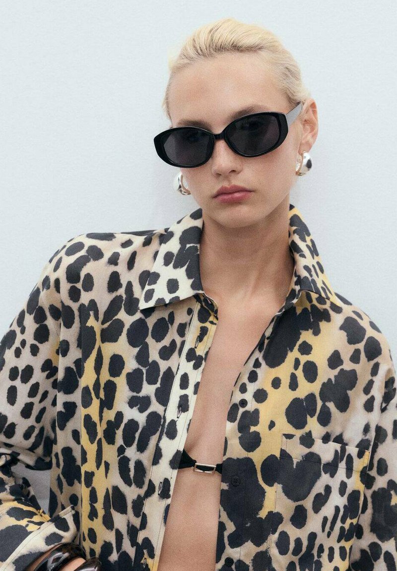 Camicia con stampa leopardata in tessuto trasparente presenta colori nero, giallo e beige, con una chiusura davanti a bottoni e una tasca sul petto. Occhiali da sole neri e orecchini audaci.