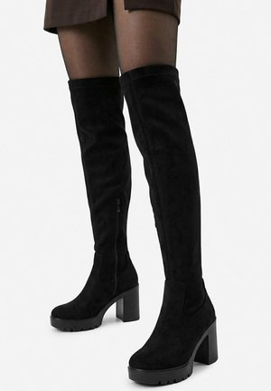 Zwarte suède over-the-knee laarzen met blokhakken en zijritsen, gedragen met donkere doorschijnende panty's en een korte rok.