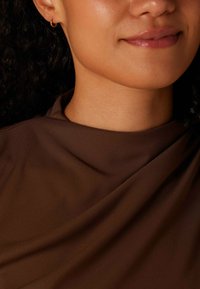 Een bruine stof hangt over het sleutelbeen, met een zachte textuur en minimalistisch ontwerp. Een kleine gouden ringoorbel is zichtbaar.