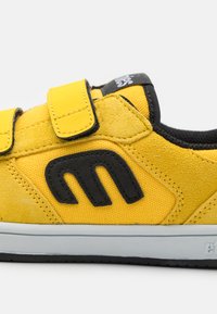 Adidași galbeni care combină pielea întoarsă și pânza, cu două curele Velcro, accente negre și o talpă din cauciuc alb.