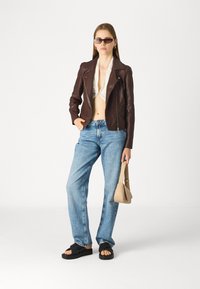 Brun skinnjakke, kremhvit topp, blå jeans, svarte sandaler og beige håndveske; modellen står mot en enkel hvit bakgrunn.