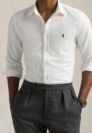 Homme portant une chemise blanche boutonnée aux manches retroussées et un pantalon plissé gris foncé, mains dans les poches, avec une montre-bracelet.
