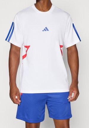 Mann trägt ein weißes Sportshirt mit kurzen Ärmeln, blauen Streifen und roten Akzenten, kombiniert mit blauen Sportshorts.