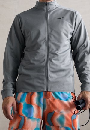 Personne portant une veste grise zippée et un short coloré aux motifs orange, bleu et blanc, tenant une corde à sauter noire dans la main droite.