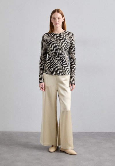 Bluza cu mâneci lungi în dungi de zebră, bej și negru, asortată cu pantaloni largi crem și pantofi bej corespunzători, pe un fond neutru.
