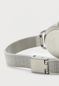 Relógio em aço inoxidável com bracelete em malha, apresentando um acabamento metálico e uma fivela gravada com o nome da marca. Mostrador redondo visível.