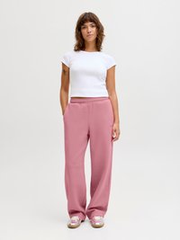 Vrouw staand met handen in de zakken, gekleed in een wit kortarmig shirt, roze joggingbroek en zilverroze sneakers tegen een effen achtergrond.