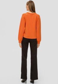 Oranje pullover met een ronde halslijn en lange, gepofte mouwen; gecombineerd met zwarte, flared broeken. De stof lijkt zacht met een gladde textuur.