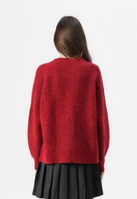Pull en maille rouge texturé, à coupe décontractée et épaules tombantes, associé à une jupe plissée noire. Pas de fermetures ou de motifs visibles.