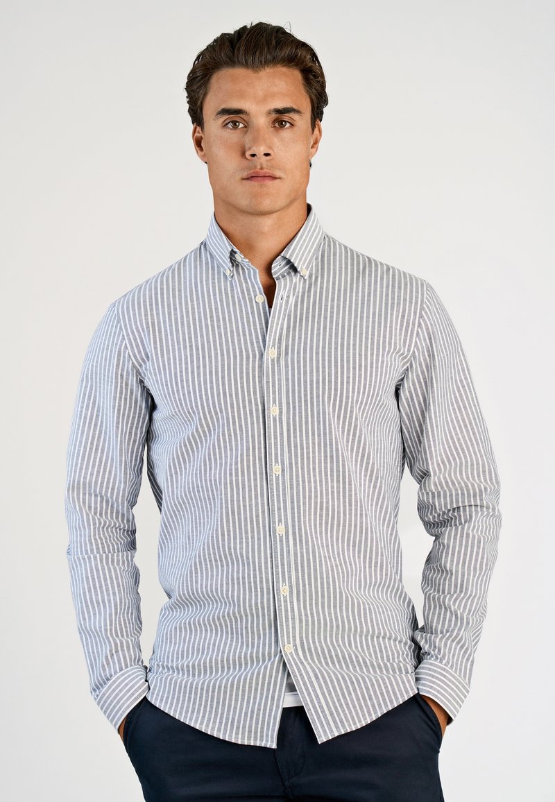 Πουκάμισο με κου μπιά που έχει κατακόρυφες ρίγες σε μπλε και λευκό, μακριά μανίκια και κλασικό γιακά. Κατασκευασμένο από ελαφρύ ύφασμα με λεία υφή.