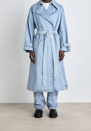 Personne portant un long trench-coat bleu à carreaux ceinturé à la taille, associé à un jean bleu clair et des chaussures noires épaisses.