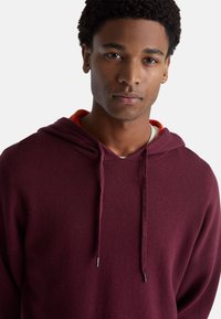 Sudadera con capucha en color marrón, hecha de una tela suave y que cuenta con un cordón con puntas de metal. Lleva una capa de acento naranja debajo.