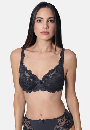 Frau trägt dunkelgrauen Spitzen-BH mit Blumenmuster und passende Unterwäsche, steht vor einfarbigem hellgrauem Hintergrund.