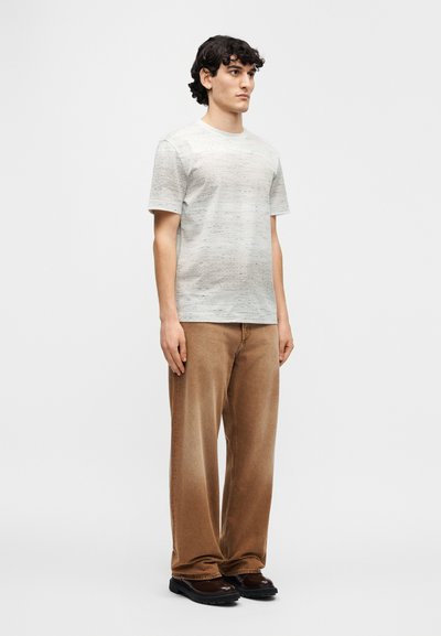 Ανοιχτό γκρι πλεκτό μπλουζάκι με υφή, σε συνδυασμό με καφέ παντελόνι φαρδύ πόδι και μαύρα παπούτσια. Πρόσοψη, σε όρθια στάση.