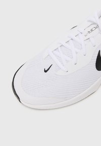 Hvid atletisk sko med en tekstureret mesh-overdel, hvide snørebånd, et sort Nike-logo og en sort gummisål med rillede detaljer.