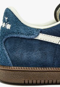 Sneaker blu scamosciato con superficie texturizzata, dettagli bianchi sui lati e suola in gomma marrone texturizzata con motivo a righe.