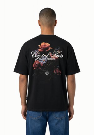 Homme portant un T-shirt noir avec un motif floral et le texte "Crystal Paris Calm But Powerful" dans le dos, associé à un jean bleu, debout de dos.
