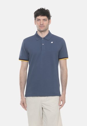 Uomo che indossa una polo blu navy con polsini a righe arancioni e gialle e pantaloni beige, in piedi davanti a uno sfondo bianco.