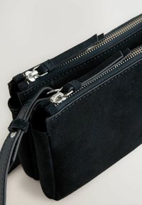 Sort ruskinds crossbody taske med dobbelt lynlåslommer, sølv hardware og sorte læderdetaljer. Glat tekstur med rene linjer.