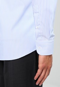 De hand van een man rustend aan de zijkant, gekleed in een lichtblauw en wit verticaal gestreept overhemd met lange mouwen en donkere broek.