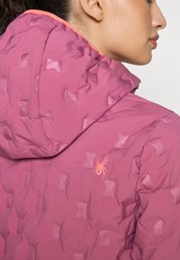 Μωβ μπουφάν με υφή, διαθέτει κουκούλα και αντίθεση πορτοκαλί λεπτομέρειες. Στο πλάι περιλαμβάνει ένα ροζ λογότυπο αράχνης.