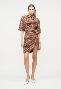 Γυναίκα ντυμένη με καφέ φόρεμα κοντομάνικο με σχέδια και ασύμμετρο τελείωμα, φορεμένα λευκά ψηλοτάκουνα παπούτσια με μυτερές μύτες, που στέκεται μπροστά σε απλό φόντο.