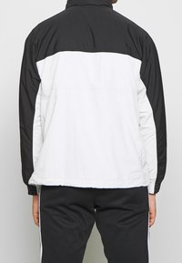 Windbreaker jacket featuring a black upper section and white lower section, with elastic cuffs and a stand collar. Material appears lightweight.