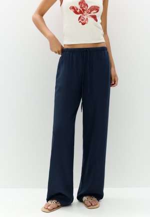 Kobieta ubrana w granatowe spodnie o szerokich nogawkach z wiązaniem w pasie, biały top na ramiączkach z czerwonym kwiatowym nadrukiem oraz beżowe sandały, stojąca na tle białego tła.