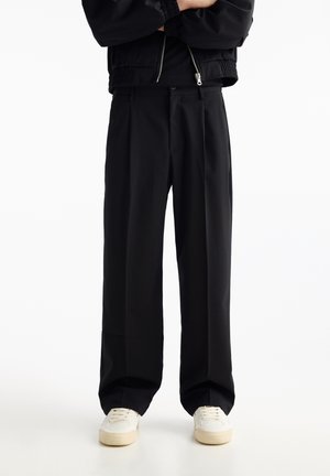 Personne portant un pantalon noir ample, des baskets blanches et une veste noire zippée, bras croisés.