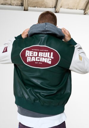 Person passt graue Kapuze an und trägt eine dunkelgrüne und weiße Jacke mit "Red Bull Racing" Aufnäher auf dem Rücken in einer neutralen Innenumgebung.