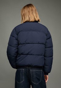 Tmavě modrá prošívaná bunda s prošívaným vzorem, vysokým límcem a elastickými manžetami, ve spojení s tmavě modrými džínami.