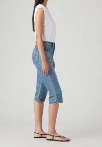 Džínové capri kalhoty ve středně modré barvě, které mají vysoký pas, kontrastní prošívání a hrubě stříhané lemy se bočními rozparky, spárované s bílým topem bez rukávů.