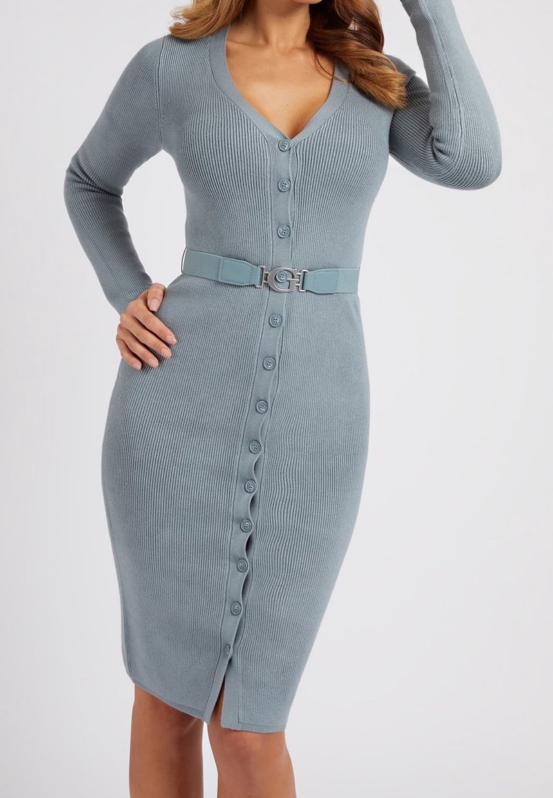 Femme portant une robe ajustée en maille côtelée bleu clair, boutonnée, avec col en V et ceinture assortie, main sur la hanche, fond neutre.