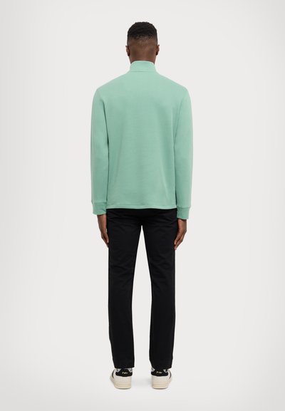 Chemise mint green à manches longues avec un col haut, assortie à un pantalon noir et des baskets blanches avec des accents noirs. Texture lisse sur l'ensemble.