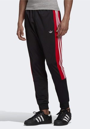 Man draagt zwarte en rode Adidas trainingsbroek met witte strepen en zwarte Adidas sneakers met witte strepen.