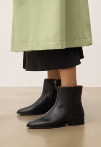 Botas de cuero negro hasta el tobillo con punteras puntiagudas y un cierre lateral con un acento dorado. Acabado mate con tacones bajos en bloque.