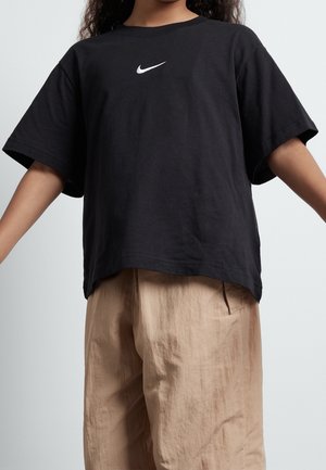 Bambino con una maglietta nera Nike larga e pantaloni beige, in piedi con le braccia leggermente estese contro uno sfondo chiaro e semplice.