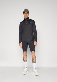 Svart, langermet genser med glidelås og høy krage, kombinert med svarte shorts. Antrekket er komplettert med hvite sneaker og en olivengrønn caps.