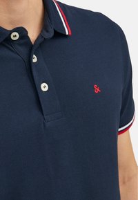 Marine poloshirt met een textuurstof, drie witte knopen en een rood geborduurd logo. Voorzien van contrasterende kraagafwerking in rood, wit en blauw.
