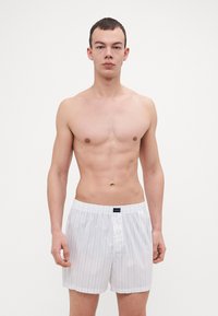 Beli bombažni bokserji z modrimi navpičnimi črtami, z gumbom za zapiranje in elastičnim pasom. Material se zdi gladek in lahek.